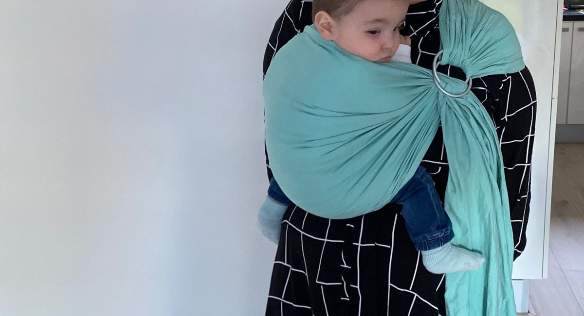 Een baby in een turquoise ring sling tegen een lichte muur